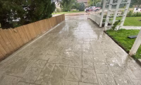 Wet stamped concrete driveway after rain, showcasing detailed tile patterns bordered by a wooden fence and green lawn, highlighting Hugo's Concrete craftsmanship in Northern Colorado.