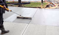 A construction worker in Northern Colorado smoothing freshly poured concrete on a new sidewalk, using a long-handled trowel under overcast conditions.