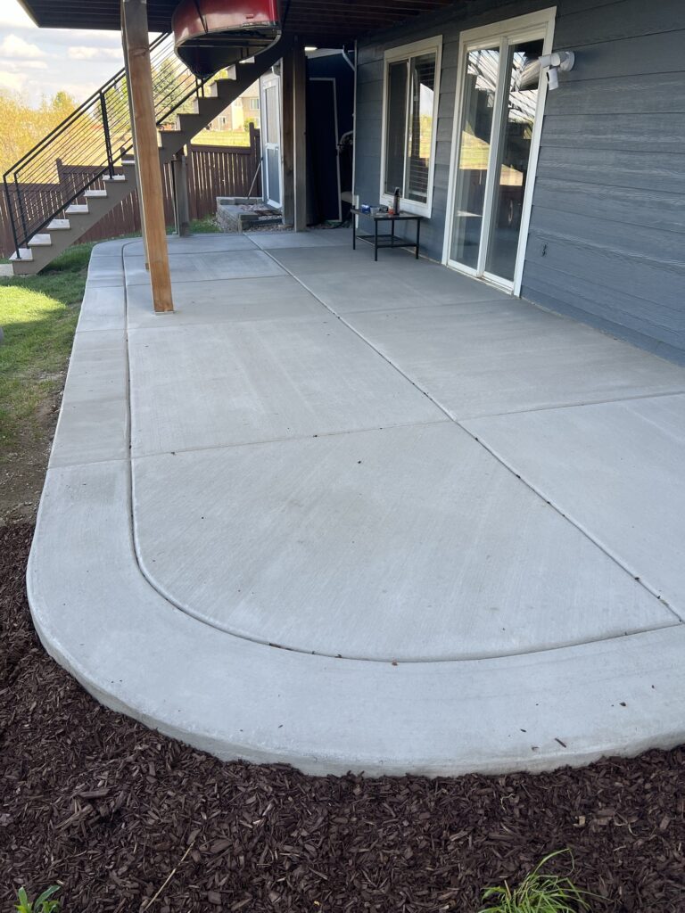 Newly installed custom concrete patio with smooth finish, featuring curved edges and a modern design under a raised deck.